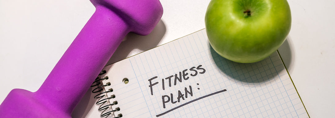 Purple dumbbell beside a green apple on a notebook with Fitness Plan written on it.